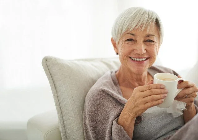 Elderly woman with grey hair smiling while holding a cup, seated on a couch.