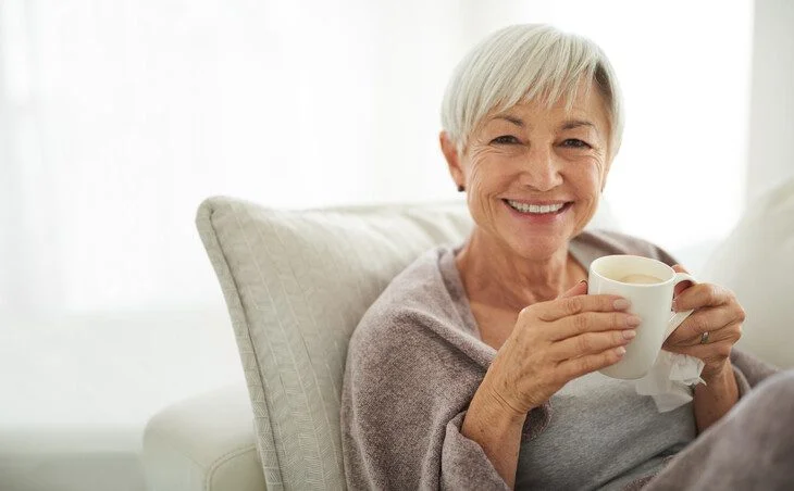Elderly woman with grey hair smiling while holding a cup, seated on a couch.
