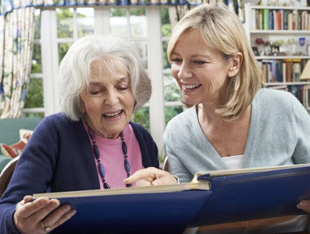 Two women smiling and looking at a photo album together in a cozy living room.