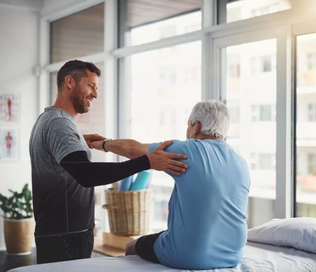 Physical therapist assists senior with arm exercises near large windows.