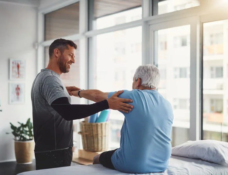 Physical therapist assists senior with arm exercises near large windows.