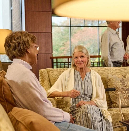 Two women seated on a couch in conversation, with two men chatting in the background.