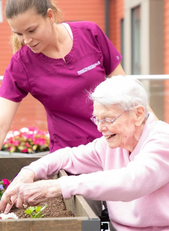 Caregiver helps elderly woman with gardening in a raised flower bed outdoors.