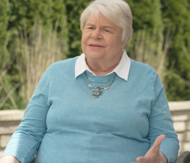 Elderly woman with short hair speaks outdoors, wearing a blue sweater and necklace.