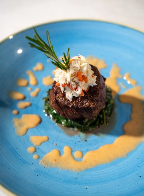Steak topped with crab and rosemary on a blue plate with sauce drizzle.