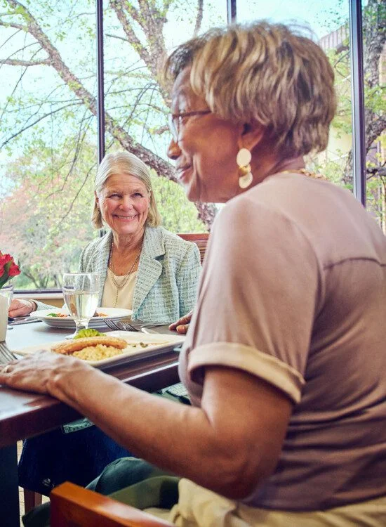 Elderly women sharing a meal and conversation in a bright dining area.