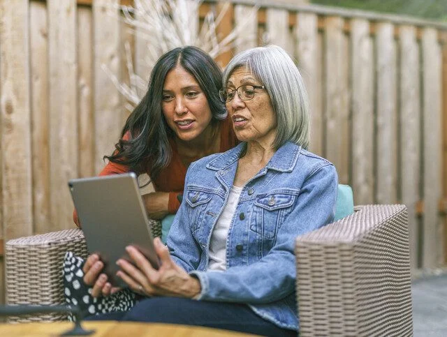 Older adult woman sitting outside while showing tablet to older adult daughter.