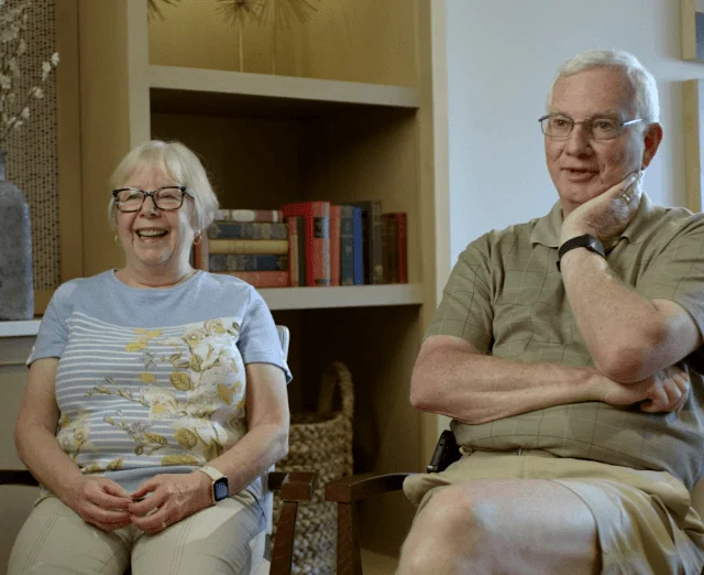 Two seniors sitting and smiling in a cozy room with bookshelves.