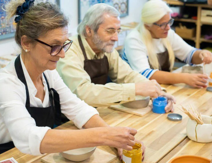 Three people painting pottery at a workshop table, focused and engaged in their work.