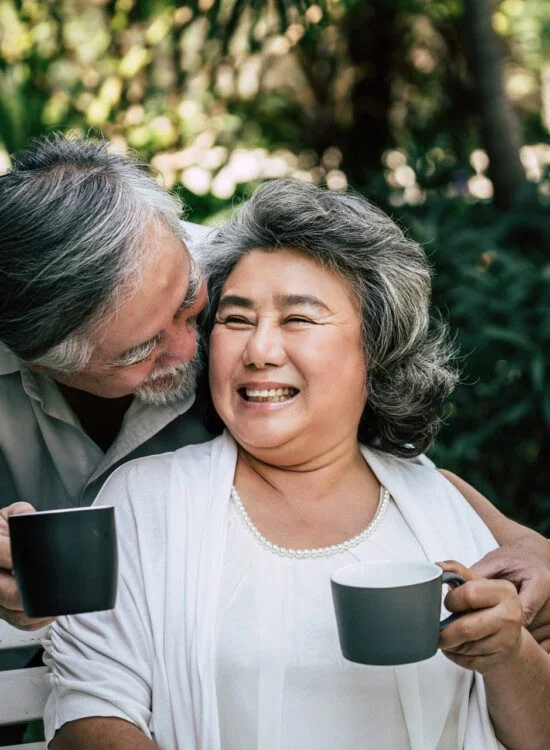 Elderly couple smiling and holding coffee mugs outdoors.