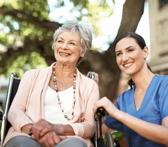 Elderly woman in wheelchair smiling with a caregiver in a garden setting.