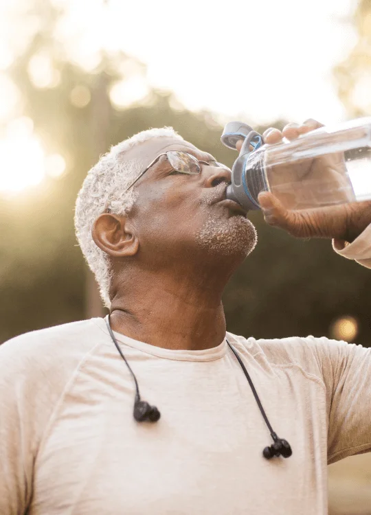 Man outdoors drinking water from a bottle, wearing earphones and sunglasses.