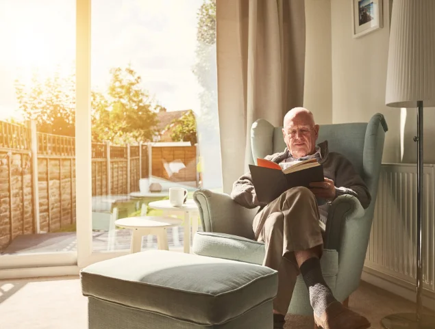 Elderly man reads a book in an armchair by a sunlit window.