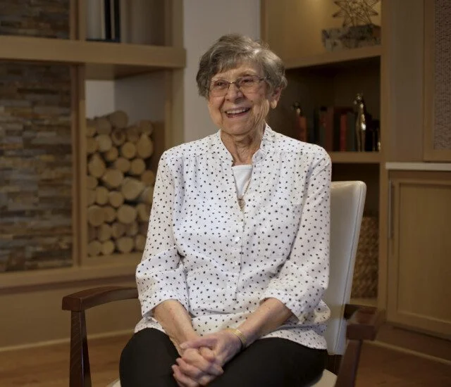 Elderly woman smiling, seated indoors with wood and bookshelves in background.