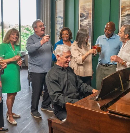 A group celebrates around a piano with drinks and smiles.