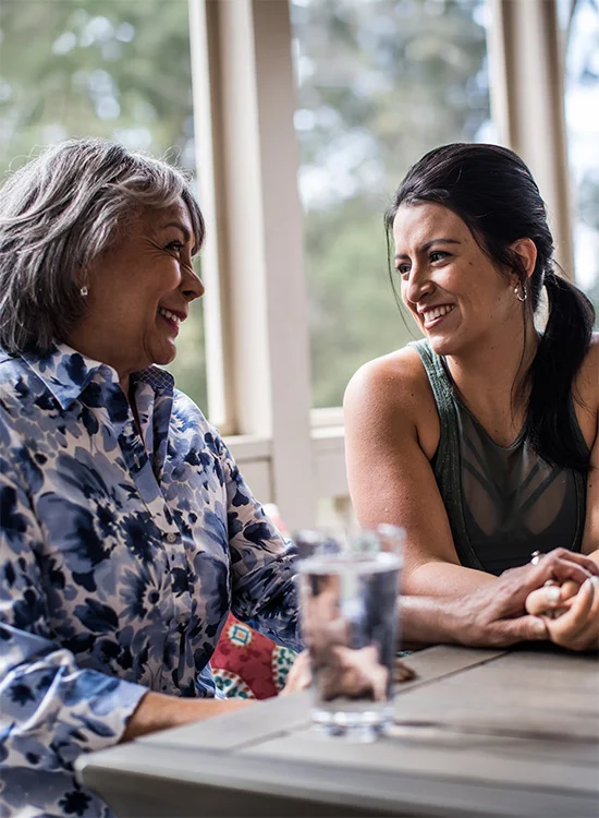 Two women share laughter at a table with water glasses and natural light behind them.