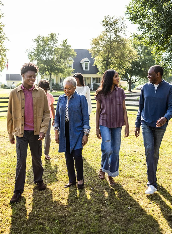 A family walks together on grass near a house and trees under sunny skies.
