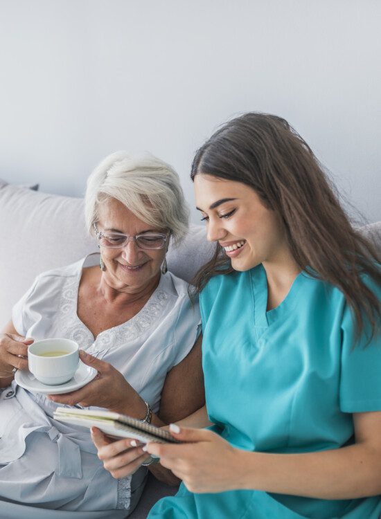 Happy patient is holding caregiver for a hand while spending time together. Elderly woman in nursing home and nurse. Aged elegant woman and tea time at nursing home