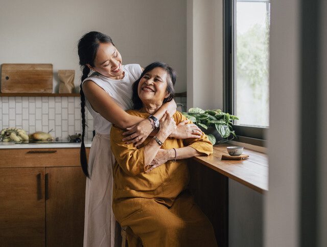 A Happy Beautiful Woman Hugging Her Mother While She Is Sitting In The Kitchen And Drinking Tea