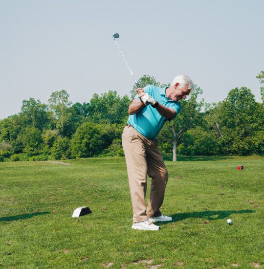Two men play golf on sunny day with trees and grassy field backdrop.